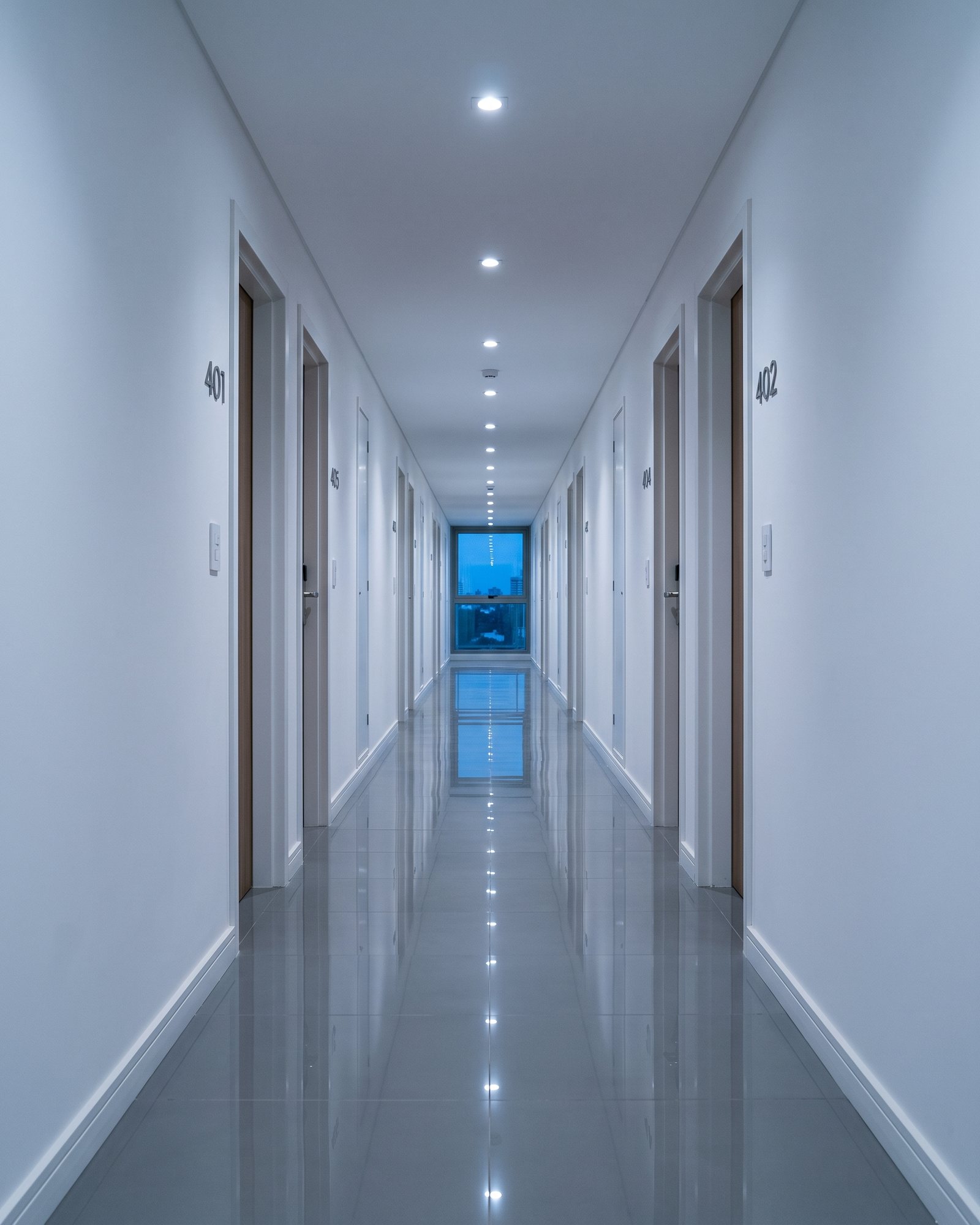 Corredor de um edifício residencial, com as portas numeradas dos apartamentos e uma janela ao fundo